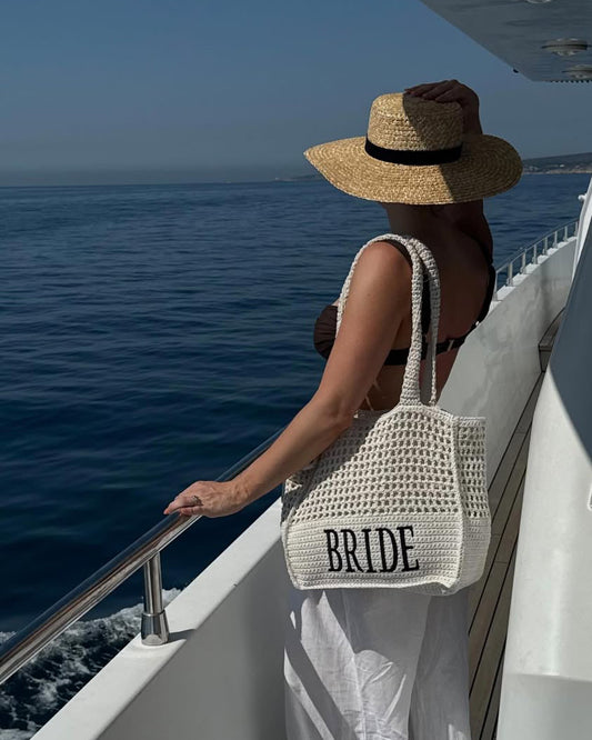Woman on a boat wearing a 'Bride' bag and straw hat, looking at the ocean.